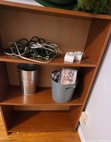 Small melamine bookshelf with a silver metal bucket, gray plastic bucket containing CDs, and electrical cords on upper shelf, photographed against a wall with electrical outlet
