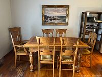 Full view of vintage oak dining table with six chairs and one captain chair arranged around it, showing wooden floor and artwork on wall.