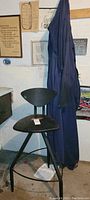 Photo of black metal swivel workshop chair with curved backrest and tripod base standing next to hanging dark blue work suits.