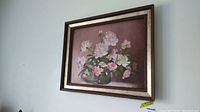 Framed floral painting on board showing pink and white flowers in a glass vase against a rose background, with gold-toned frame and cream inner border.