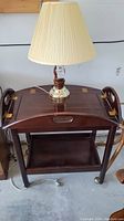 Wooden bar cart with removable tray on top and lower shelf, holding a table lamp with beige pleated lampshade. Cart has caster wheels.