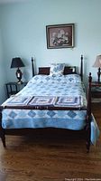 Full view of vintage wooden double bed with blue and white bedding, decorative crochet squares folded at foot of bed, wood floor.