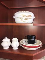 Several ceramic items including sugar bowl with lid, creamer, small plates, and a black coffee mug on a wooden shelf.