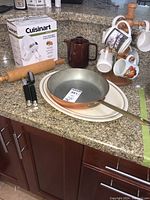Photo of countertop with boxed Cuisinart hand mixer, mugs on white metal stand, metal frying pan with wooden handle, brown ceramic teapot, rolling pin, large plates, and hand can opener.