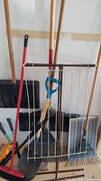 Photo showing various garden tools leaning against a wall including a cloth drying rack, shovels, brooms, rakes, and hedge shears.