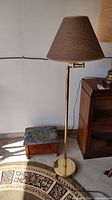 View of floor lamp with brass finish and swivel arm next to wood footstool with floral top, showing overall size and design