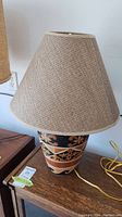 Full view of the lamp showing woven fabric lampshade and ceramic base with painted geometric and floral motifs in black, orange, beige.