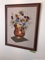 Frontal view of the larger framed needlepoint showing the floral bouquet with brown vase and multicolored flowers on beige fabric, framed in dark wood.