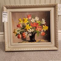 Framed oil on board painting showing a flower arrangement in a black vase with tulips, daffodils and other flowers on a neutral background.