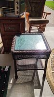 Square faux wicker table with glass top and lower woven shelf, shown in natural light among other furniture items.