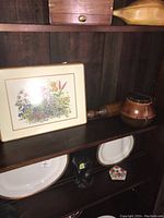 Shelf display showing one leather covered lidded jar, a wooden rolling pin, and a floral placemat mounted on board, along with some other items not included in lot