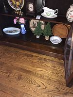 Image showing the lower shelf of a dark wood cabinet containing the Royal Worcester oval dish, blue and white vase with pink flowers, two small green artificial Christmas trees, two wooden serving boards, and two small floral bowls.