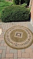 Round outdoor rug placed on stone pavement bordered by shrubbery, showing full rug design and surroundings.