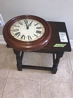 Round wood frame wall clock with Roman numeral face and battery operated quartz mechanism placed on stool.