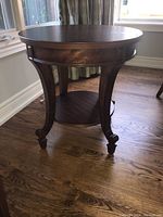Full side view of the wooden round side table showing curved legs and lower shelf on hardwood floor.