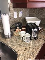 Kitchen countertop with Black and Decker toaster, four mugs, small electric fan, white plastic bin, and decorative items near kitchen sink