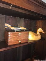 Wooden duck decoy, wooden desk box with metal corner accents and brass knob, plastic jet fighter model on dark wood shelf.