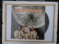 Front cover of Apollo 11 vinyl record showing astronauts and the moon with title 'We Have Landed on the Moon'. The vinyl is partially visible inside the sleeve.