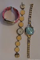 Photo showing three women's fashion watches: a pink silicone strap watch with a rectangular face, a silver-tone bracelet watch with beige round discs, and a silver metal band watch with a blue face labeled XOXO.