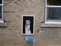 Framed painting of a woman in a blue bonnet holding an orange flower against a black background, leaning on a chair outside near a brick wall.