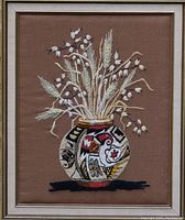 Close-up photo of the woven textile art featuring Native American style pottery and floral elements framed and mounted on brown fabric background.