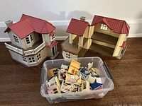 Two Calico Critters dollhouses, one closed and one open to show interior, with a plastic container in front filled with miniature furniture pieces and animal figurines.