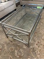Photo of rectangular coffee table with glass top and metal frame showing crossbar detailing and wheels, placed on carpet in living room.