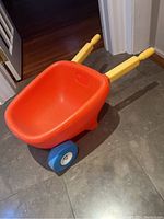 Photo of the front and side view of the child's wheelbarrow showing red tub, yellow handles, and blue single wheel on tile floor.