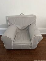 Front view of grey kids chair with white piping, showing padded arms, back, and seat with removable cover. Fabric handle is visible at top rear.