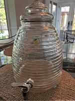 Clear ribbed glass water dispenser sitting on a table with a matching lid and a chrome spigot near the base. The dispenser has a small circular sticker on it.