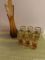 Full lot showing tall amber glass vase with scalloped edges and six matching mini liquor glasses in amber glass with gold leaf accents, displayed on fabric surface