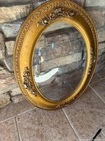 Full front view of the antique oval frame with gold finish and convex glass positioned upright against a stone wall.