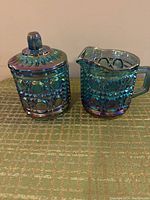 Front view of the blue carnival glass sugar bowl with lid and cream pitcher, showing the iridescent pattern and opalescent blue finish.