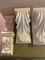 Four white ceramic ornate wall shelves, showing large leaf motif shelves and smaller acanthus leaf carved shelves on green fabric.