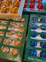 Multiple sets of gold, pearl-white, red, and blue spherical glass Christmas ornaments still in original green packaging boxes labeled 'Holiday Classics' and '20 Traditional Glass Ornaments'.