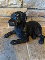 Side view of the heavy concrete black painted dog statue on tile floor against stone wall background.