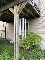 Wind chime hanging from a wooden post beside a garden, showing five white metal tubular chimes hanging from a weathered wooden frame.