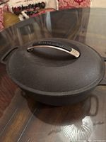 Closed black cast iron pot with a silver KitchenAid branded handle on the lid, placed on a glass table.