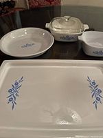 Display of all four Corning Ware pieces showing blue cornflower floral pattern on white ceramic surface: broil/bake tray, pie plate, two baking dishes with glass lid visible.