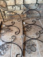 Six black metal hanging hooks with decorative bee, frog, and butterfly wire-frame shapes arranged on tile floor against stone wall background.