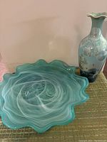 Wide view of the aqua blue scalloped glass platter with white swirls next to the tall floral glass vase