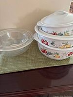Photo displaying three clear nested Pyrex glass bowls alongside three white Corelle heavy bowls with colorful fruit patterns and lids on a table with a green mat.