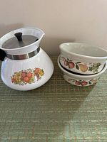 Photo of white ceramic Corning Ware P104 teapot with vintage decorative fruit design and black handle and knob, alongside two stacked Wedgewood bowls showing quince fruit pattern.