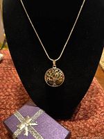 Full view of the Tree of Life pendant necklace displayed on a black stand alongside a small purple gift box wrapped with a silver ribbon.
