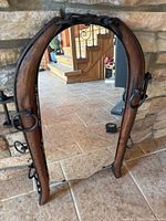 Full view of a horse yoke shaped mirror standing on a tiled floor in front of a stone wall and rustic wooden stairs in background.
