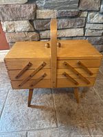 Closed accordion-style wooden sewing basket with carrying handle, showing light wood and dovetail joints.