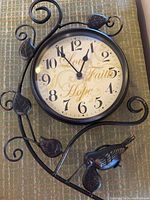 Full view of the black metal wall clock showing round face with black numerals and decorative bird and leaves on metal frame.