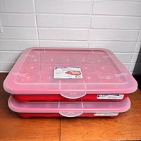 Two closed plastic storage containers stacked, showing translucent pinkish lids with snap closures and red compartment base.