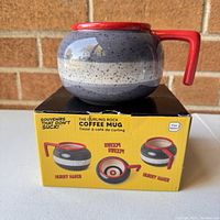 Photo of curling stone coffee mug displayed on box, showing shape and colors including speckled gray stone body, cream band, and red handle.