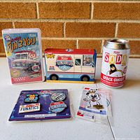 Front view of Funko Freddy Funko figure, Fun Run ice cream truck figure, Funko Soda Space Ghost can, collectible pins, and small vinyl figure keychain on a white surface against a brick background.